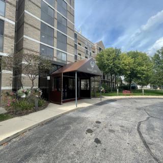 The image shows the entrance of a multi-story building with a canopy, surrounded by trees and a paved driveway under a partly cloudy sky.