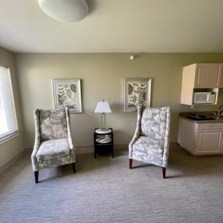 A minimalist living room featuring two patterned chairs, a small side table with a lamp, artwork on the walls, and a kitchenette in the corner.