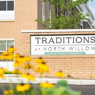 A sign for Traditions at North Willow in front of a building, with yellow flowers in the foreground.
