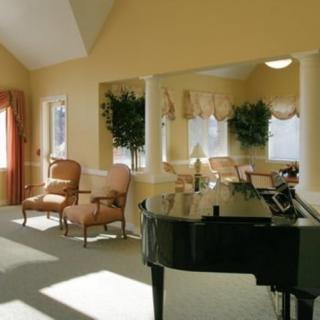 A cozy living room with a grand piano, elegantly furnished with chairs and greenery, bathed in natural light.