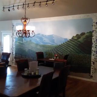 A dining room with a vineyard mural covering one wall, a chandelier hanging from the ceiling, and a wooden table surrounded by chairs.