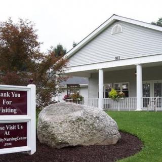 A white building with a porch, surrounded by greenery and featuring a prominent rock in front with a sign nearby.