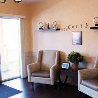 A cozy waiting room with two armchairs, decorative items on shelves, and natural light coming through large windows.