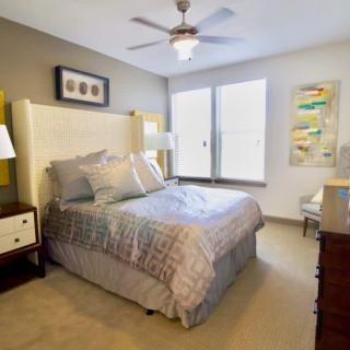 A cozy and well-decorated bedroom featuring a bed with patterned bedding, bedside tables, lamps, artwork on the walls, and a ceiling fan.