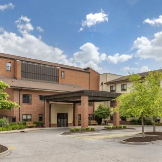 A hotel building with a brick exterior and a covered entrance, surrounded by trees and a parking area under a clear blue sky.