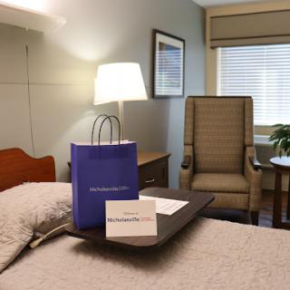 A cozy hotel room featuring a bed with a decorative lamp, a chair for relaxation, and a gift bag on the table.