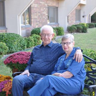 An elderly couple sitting together on a bench in a garden setting.