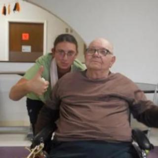 An elderly man in a wheelchair with a caretaker giving a thumbs up.