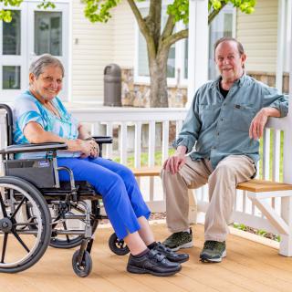 An elderly woman in a wheelchair sitting next to a man on a bench, enjoying time outdoors.