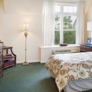 A cozy bedroom with a large window, featuring a bed with patterned bedding, a standing lamp, and wooden furniture.