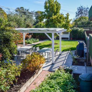 A tranquil backyard features a white pergola with outdoor seating, surrounded by lush greenery and garden on a sunny day.