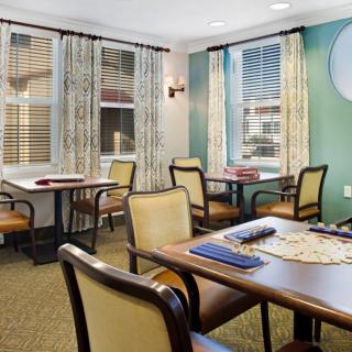 A cozy game room with tables and chairs, featuring windows with curtains and board games on the tables.
