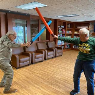 Two seniors engage in a playful sword fight with foam swords in a cozy room.