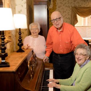 A group of seniors enjoying time together around a piano in a cozy living room.