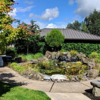 A scenic garden with a pond surrounded by lush greenery and trees under a partly cloudy sky.