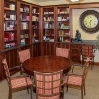A cozy library corner with a round wooden table surrounded by chairs, bookshelves filled with books, and a large wall clock.