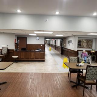 A spacious common area in a senior living facility, featuring tables, chairs, and a hallway leading to other areas.