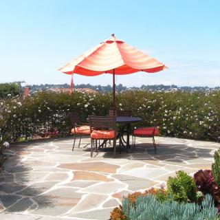 A sunny patio with a table and chairs under an orange umbrella, surrounded by a garden.