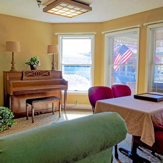 A cozy living room featuring a piano, green couch, and a table with red chairs overlooking an American flag outside the window.