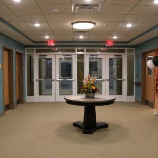 A well-lit lobby featuring a central table with floral decor, surrounded by glass doors and neutral-colored walls.