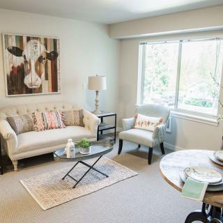 A cozy living room featuring a couch, armchair, coffee table, and a large window. The walls are adorned with artwork that adds character to the space.