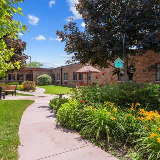 A scenic garden pathway surrounded by lush greenery, flowers, and trees, leading to a brick building under a bright blue sky.