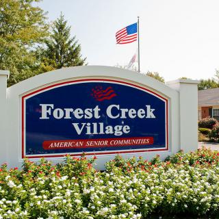 Entrance sign for Forest Creek Village, an American Senior Community, with a flag and landscaped flowers.