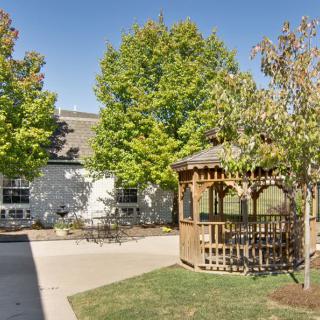 A peaceful outdoor scene featuring a wooden gazebo surrounded by trees and a building.