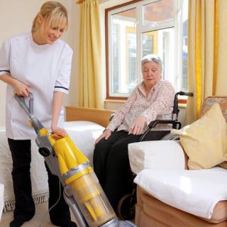 A caregiver vacuuming the room while assisting an elderly person.