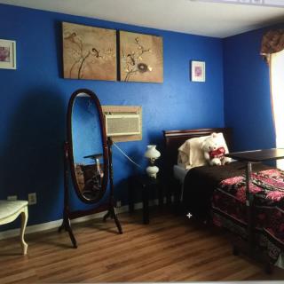 A cozy bedroom with blue walls, featuring a decorative mirror, artwork, and natural light streaming through the window.