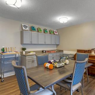 A cozy dining area with a table, chairs, and nearby art supplies, highlighted by a piano and shelving for decor.