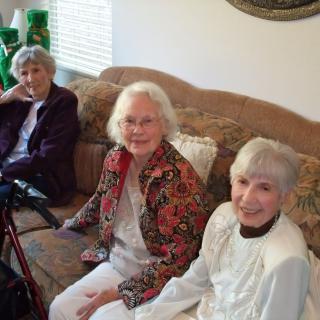 A group of senior women sitting together in a living room, smiling at the camera.