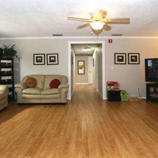 A cozy living room with hardwood flooring, featuring a sofa, television, and ceiling fan, leading into a hallway.