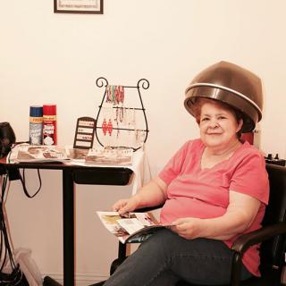 A woman sits under a hair dryer in a salon, reading a magazine.