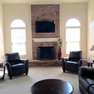 A cozy living room featuring a brick fireplace and a mounted television, flanked by two large windows and surrounded by comfortable armchairs.