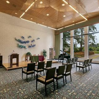 A spacious room designed for gatherings, featuring a wooden ceiling and natural lighting, with chairs arranged in front of a small stage.