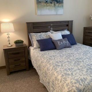 A cozy bedroom with a neatly made bed, adorned with decorative pillows and a nightstand featuring a lamp.