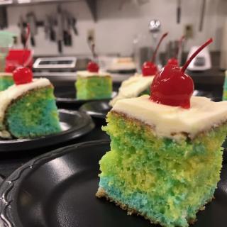 A slice of colorful cake topped with creamy frosting and a cherry.