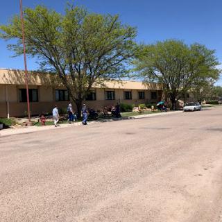 A view of a street lined with trees and a building, featuring people walking along the sidewalk.