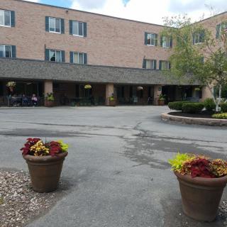 A three-story brick building with a main entrance surrounded by potted plants and a parking area.