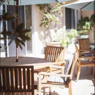A sunlit patio featuring wooden chairs and tables under umbrellas, surrounded by greenery.
