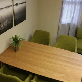 A small meeting room with a wooden table surrounded by green chairs, featuring a potted plant and artwork on the walls.