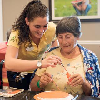 A caregiver assists a senior woman in a painting activity.
