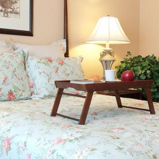 A cozy bedroom scene with floral bedding and a wooden tray on the bed, holding a red apple and a cup, beside a decorative lamp.