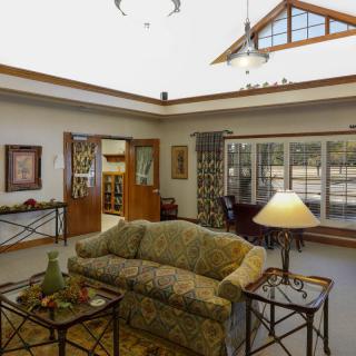 A cozy living room with a couch, tables, and a lamp, illuminated by natural light from large windows.