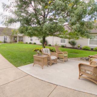 A serene outdoor patio with wicker chairs set on a concrete path surrounded by lush grass and trees.