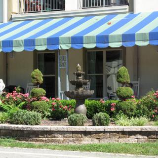 A charming garden with a striped blue and green awning, featuring a central fountain surrounded by topiary shrubs and colorful flowers.