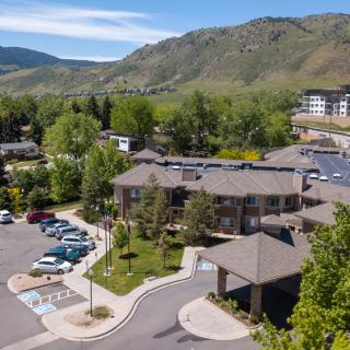 Aerial view of a building surrounded by a parking lot and lush greenery, set against a mountain backdrop on a sunny day.