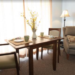 A cozy dining area with a wooden table and chairs, decorated with a vase of flowers and accompanied by a comfortable armchair and standing lamp.