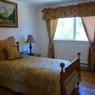 A cozy bedroom with a single bed, adorned with matching pillows and bedding, next to a nightstand with a lamp. The window is dressed with light-colored curtains and natural light fills the room, complementing the warm tones of the wooden floor.